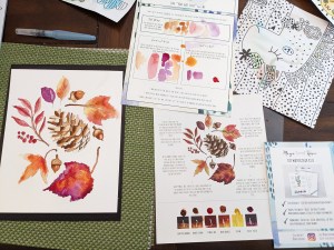 A watercolor kit - a sheet with numerous leaves encircling a pinecone and acorns. It has shades of brown and orange and red. To the right is a test sheet for colors and an instruction sheet with small dabs of paint - the source of paint for the painting.